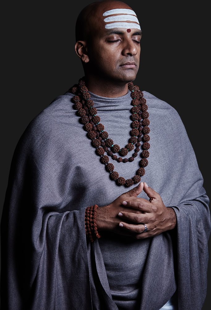 Dandapani standing with his eyes closed and hands folded, in a grey shawl and rudraksha beads.